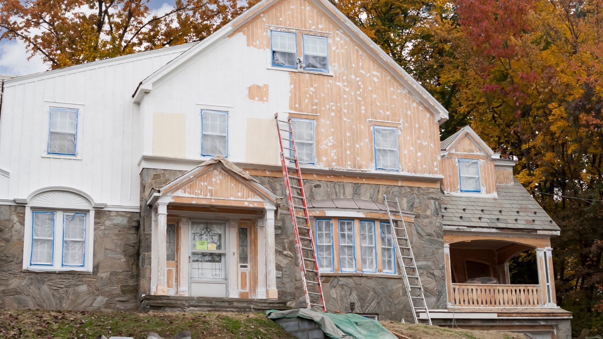Multi-story house under renovation with ladder, autumn trees in background